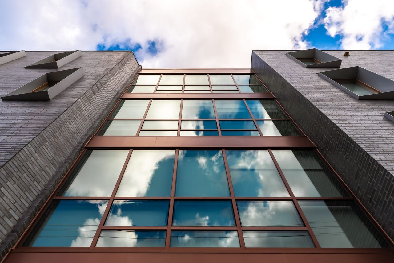 Looking up a bronze aluminum curtain wall between dark brick walls, reflecting clouds
